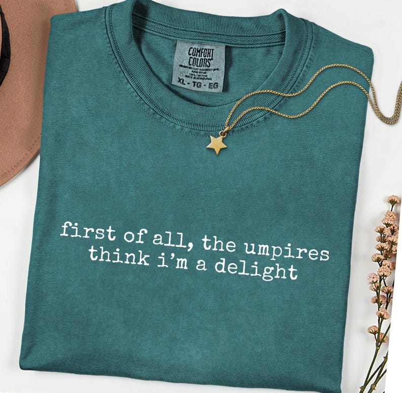 May include: Teal Comfort Colors t-shirt with the text "first of all, the umpires think i'm a delight" in white. A gold star pendant necklace rests on the shirt. The shirt has a crew neck and a tag that says XL-TG-EG.