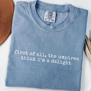 May include: A blue Comfort Colors t-shirt with the text "first of all, the umpires think i'm a delight" in white. The shirt has a crew neck and short sleeves. The tag reads "S P CH".