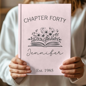 May include: A pink book with the text "CHAPTER FORTY" at the top and "Jennifer" in cursive below a floral illustration. The bottom of the book reads "Est. 1985".
