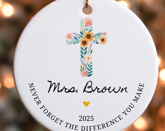 May include: White ceramic ornament with a floral cross design featuring sunflowers and other flowers. The ornament has the text "Mrs. Brown", a yellow heart, and "2025". The text "NEVER FORGET THE DIFFERENCE YOU MAKE" is also included.