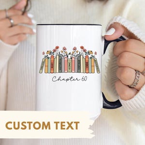 May include: White ceramic mug with a black interior and handle. The mug features a design of colorful books with floral accents and the text "Chapter 60". The words "Timeless", "Mary", "Historical", "Iconic", "Vintage", "1966", and "Masterpiece" are printed on the books.