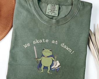 May include: An olive green crew neck t-shirt featuring a graphic of a frog wearing a purple cape, holding a sword in one hand and a pair of white ice skates in the other. The text "We skate at dawn!" is printed in white above the frog illustration.