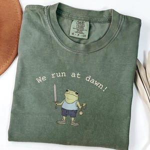 May include: Olive green t-shirt with the text "we run at dawn!" and a cartoon frog holding a sword and pocket watch. The frog wears a blue tank top, shorts, and sneakers. The shirt has a Comfort Colors tag.