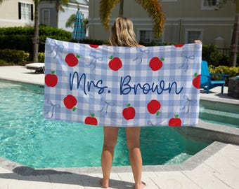 Puede incluir: Una toalla de playa a cuadros azules y blancos con diseños de manzanas rojas y lazos. La toalla tiene el texto "Mrs. Brown" en una fuente cursiva. La toalla se sostiene cerca de una piscina.