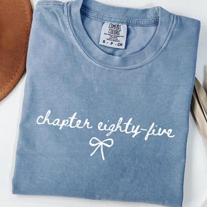 May include: A blue Comfort Colors t-shirt with the text "chapter eighty-five" in white script and a bow graphic. The shirt is folded and has a tag at the neckline indicating size.