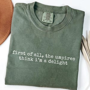 May include: Olive green t-shirt with the text "first of all, the umpires think i'm a delight" in white. The shirt has a crew neck and a Comfort Colors tag. A brown hat is partially visible in the upper left corner.