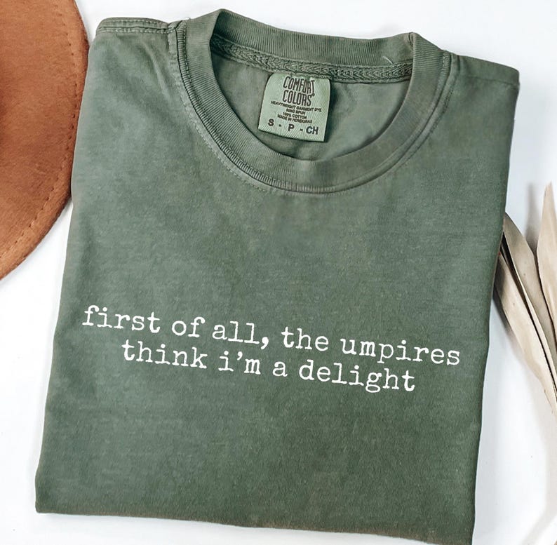 May include: Olive green t-shirt with the text "first of all, the umpires think i'm a delight" in white. The shirt has a crew neck and a Comfort Colors tag. A brown hat is partially visible in the upper left corner.