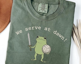 Puede incluir: Una camiseta de cuello redondo color verde oliva con un gráfico de una rana sosteniendo una espada y un balón de voleibol. El texto "We serve at dawn!" está impreso en arco sobre la rana. La camiseta tiene una etiqueta Comfort Colors en el cuello.