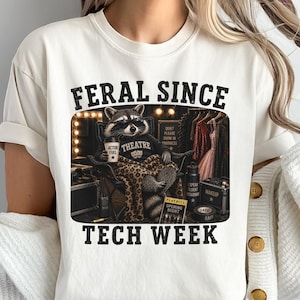 May include: A cream-colored t-shirt featuring a graphic of a raccoon wearing sunglasses and a sweater that says THEATRE, sitting in a chair with a leopard print blanket. The text FERAL SINCE TECH WEEK is printed above and below the graphic.