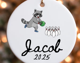 May include: White ceramic Christmas ornament with a cartoon raccoon bowling. The raccoon is gray and black, holding a green bowling ball. The ornament has the name "Jacob" and the year "2025" in black script.