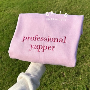 May include: A pink sweatshirt with the text "BALI'S EMBROIDERY" and "professional yapper" embroidered in pink.