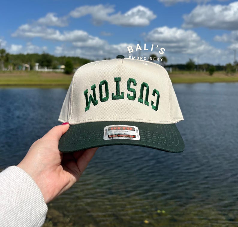 Puede incluir: Una gorra de b&eacute;isbol blanca con una visera verde y la palabra "CUSTOM" bordada en verde en la parte delantera. La gorra se sostiene en una mano contra un fondo de un lago y el cielo.