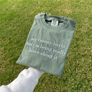 Puede incluir: Una camiseta verde con el texto "my tummy hurts but I'm being really brave about it" bordado en blanco.