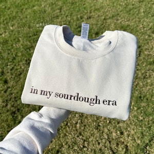 May include: A light beige sweatshirt with the text "in my sourdough era" embroidered in brown on the front.