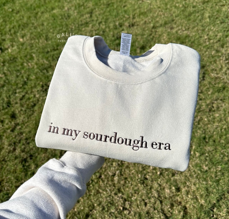 May include: A light beige sweatshirt with the text "in my sourdough era" embroidered in brown on the front.