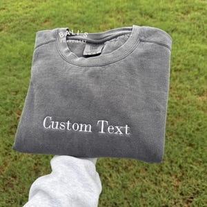 Op de afbeelding: Een antracietgrijze sweater met ronde hals en een verwassen afwerking, voorzien van de wit geborduurde tekst "Custom Text" op de borst. Het kledingstuk heeft een geribbelde kraag en een zichtbaar merklabel aan de binnenzijde "Comfort Colors".