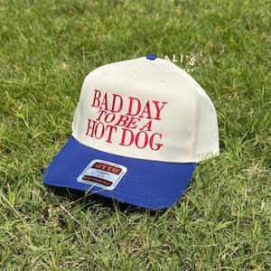 May include: A white and blue baseball cap with the text "Bad Day To Be A Hot Dog" embroidered in red on the front.