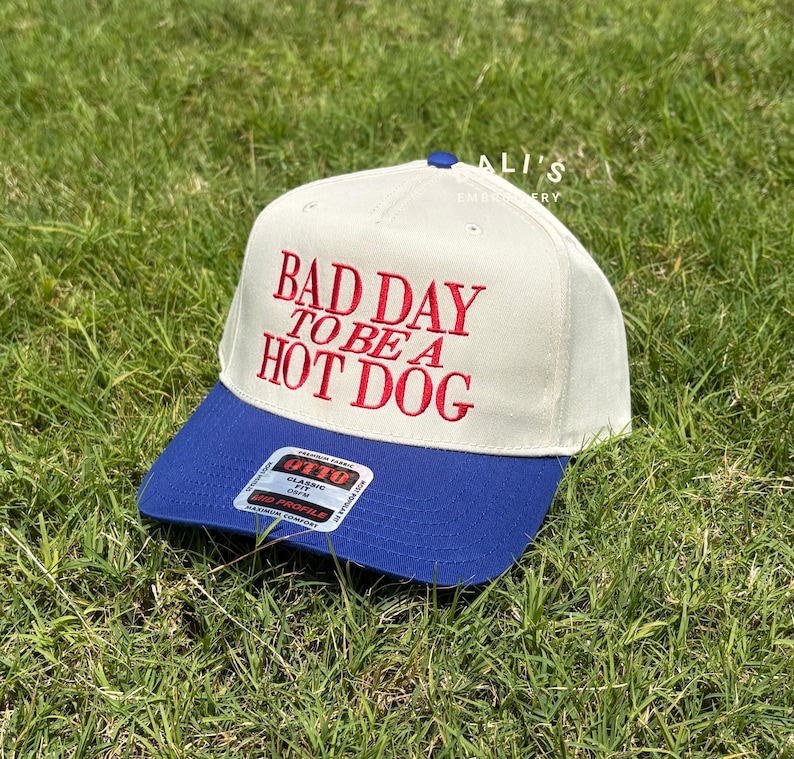 May include: A white and blue baseball cap with the text "Bad Day To Be A Hot Dog" embroidered in red on the front.