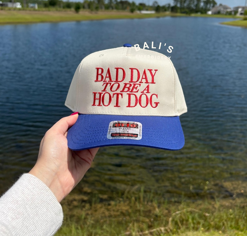 May include: A white baseball cap with a blue brim. The front of the cap has red embroidered text that reads "Bad Day To Be A Hot Dog".