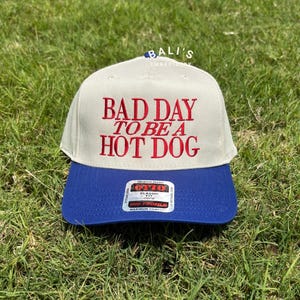 May include: A beige and blue baseball cap with the text "BAD DAY TO BE A HOT DOG" embroidered in red on the front.