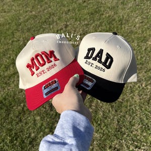 May include: Two baseball caps with the text "MOM" and "DAD" embroidered in bold letters. The "MOM" cap has a red brim and the "DAD" cap has a black brim. Both caps have "EST. 2026" embroidered below the main text.