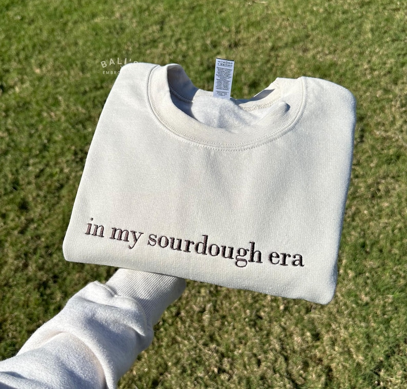 May include: A light beige sweatshirt with the text "in my sourdough era" embroidered in brown on the front.