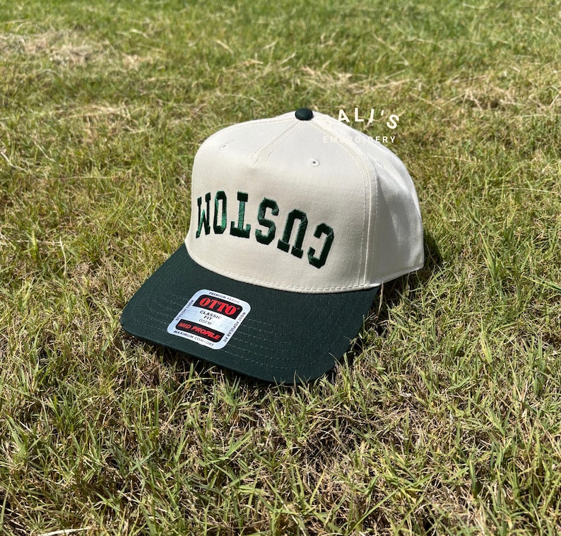 Puede incluir: Una gorra de b&eacute;isbol blanca y verde con la palabra "CUSTOM" bordada en verde en la parte delantera. La gorra tiene una visera verde y una etiqueta blanca con el texto "OTTO CLASSIC FIT" en el lateral.