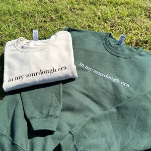 May include: Two sweatshirts, one in a cream color and one in a dark green color, both with the text "in my sourdough era" printed on them.