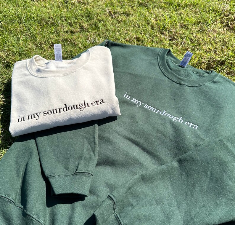 May include: Two sweatshirts, one in a cream color and one in a dark green color, both with the text "in my sourdough era" printed on them.