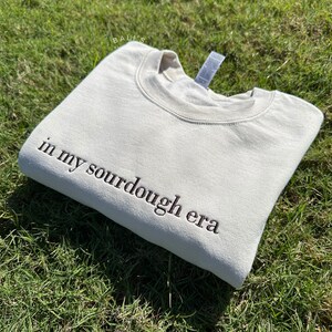 May include: A beige sweatshirt with the text "in my sourdough era" embroidered in brown on the left side of the garment.