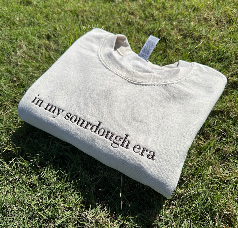 May include: A beige sweatshirt with the text "in my sourdough era" embroidered in brown on the left side of the garment.