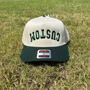 Puede incluir: Una gorra de b&eacute;isbol blanca con una visera verde y la palabra "CUSTOM" bordada en verde en la parte delantera. La gorra tiene una etiqueta en la parte posterior que dice "OTTO CLASSIC FIT 59CM MID PROFILE MAXIMUM COMFORT".