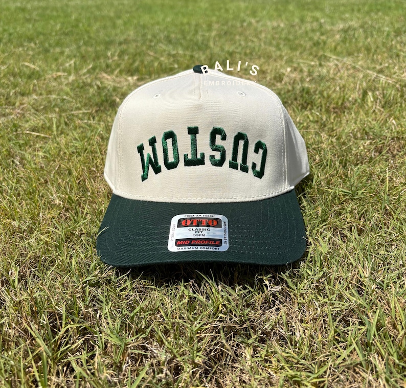 Puede incluir: Una gorra de b&eacute;isbol blanca con una visera verde y la palabra "CUSTOM" bordada en verde en la parte delantera. La gorra tiene una etiqueta en la parte posterior que dice "OTTO CLASSIC FIT 59CM MID PROFILE MAXIMUM COMFORT".