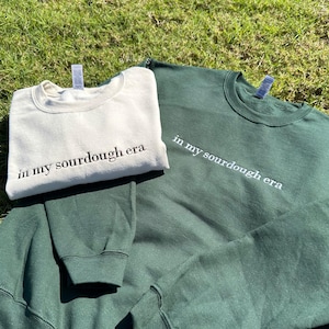 May include: Two sweatshirts, one in a cream color and one in a dark green color, both with the text "in my sourdough era" embroidered on them.