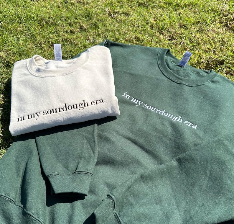 May include: Two sweatshirts, one in a cream color and one in a dark green color, both with the text "in my sourdough era" embroidered on them.