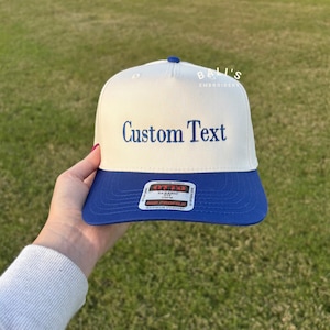 May include: A white and blue baseball cap with a blue brim. The cap has the text "Custom Text" embroidered on the front in blue thread. The cap also has a small label on the side that says "Bali's Embroidery".