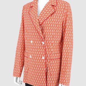 May include: A double-breasted blazer featuring a repeating geometric pattern in orange and off-white. The jacket includes a notched lapel with red piping, long sleeves, two front pockets with red trim, and four white buttons on the front closure.