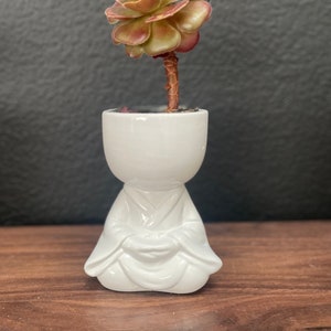 May include: A white ceramic planter shaped like a meditating person with a succulent plant growing out of the top. The succulent has green and red leaves.