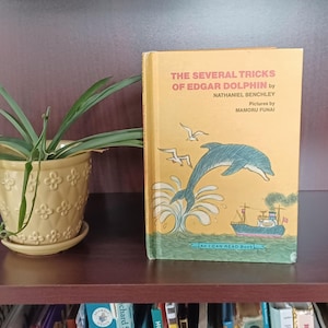 May include: A yellow book with a blue dolphin jumping out of the water. The book title is "The Several Tricks of Edgar Dolphin" by Nathaniel Benchley. The book is part of the "I Can Read Book" series.
