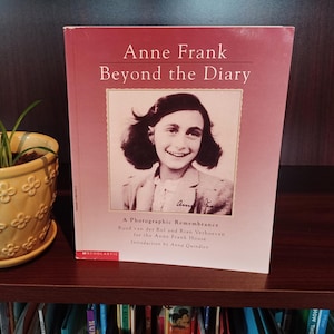 May include: A book titled "Anne Frank Beyond the Diary" with a portrait of Anne Frank on the cover. A small yellow flower pot with a plant sits to the left of the book. The book is on a wooden shelf.