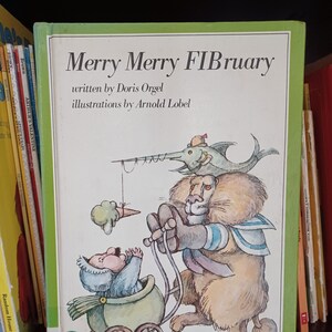 May include: A green book cover with the title "Merry Merry FIBruary" written in black. The book is illustrated with a lion wearing a sailor shirt pushing a baby in a stroller. The baby is a small, brown, furry creature with a blue hat. A blue fish with a yellow hat is perched on the lion's head.