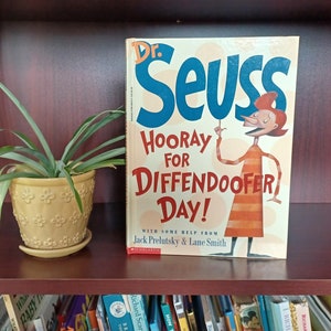May include: A colorful children's book titled "Hooray for Diffendoofer Day!" by Dr. Seuss with illustrations of a cartoon character wearing a striped shirt and a red hat. The book is on a bookshelf with other books.