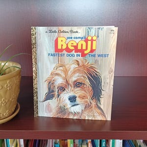 May include: A Little Golden Book titled "Benji" featuring a dog illustration. The book cover has the text "FASTEST DOG IN THE WEST" and "joe camp's". The book is displayed on a wooden shelf next to a yellow flower pot.
