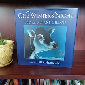 May include: A hardcover book titled "One Winter's Night" by Leo and Diane Dillon. The cover features a black and white cow illustration against a blue starry night background. The author's name is John Herman.