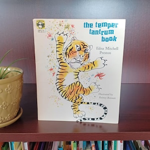 May include: A vintage children's book titled "the temper tantrum book" with a cartoon tiger illustration. The tiger is orange and black with a fierce expression. The book is on a dark brown shelf next to a yellow flower pot.