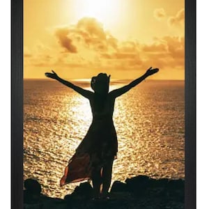 May include: Silhouette of a person with arms outstretched standing on a rocky cliff overlooking a body of water at sunset. The sky is a vibrant orange and yellow.