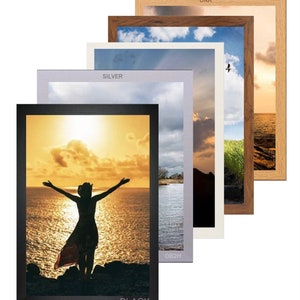 May include: Five picture frames in different colours, including black, silver, oak, and white. The frames are stacked on top of each other, with the black frame at the bottom and the oak frame at the top. The frames are all the same size and have a simple, modern design.