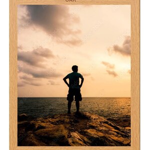 May include: A silhouette of a person standing on a rocky cliff overlooking a body of water at sunset. The sky is a vibrant orange and pink, with clouds scattered across the horizon.