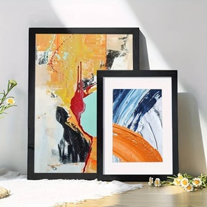 May include: Two black framed abstract paintings with colourful brushstrokes. The larger painting features a mix of yellow, orange, red, black, and white. The smaller painting features blue, white, and orange brushstrokes.