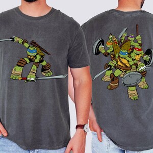 May include: A charcoal gray short-sleeve t-shirt featuring a graphic of Leonardo from the Teenage Mutant Ninja Turtles on the front and a group illustration of all four Teenage Mutant Ninja Turtles on the back, each with their signature weapons.
